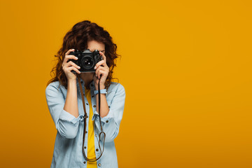 curly redhead photographer covering face with digital camera isolated on orange - obrazy, fototapety, plakaty