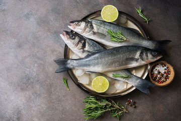 Flat lay fresh raw fish seabass in a plate with ice cubes, rosemary and lime on a dark rustic background. Top view, copy space. - obrazy, fototapety, plakaty