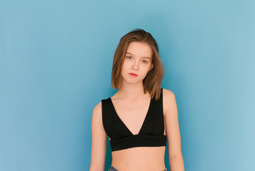 Attractive girl with a light make-up wears a bra, standing on a blue background, looking into the camera with a serious face. Model tests in agence. Model's snapshots. Snaps for agency.