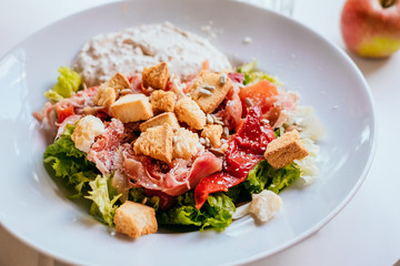 Plate of salad with tomatoes, jamon and baked vegetables on the white table. Glass of iced water with mint and lemon.