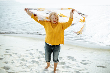 Senior woman in bright sweater enjoying sea breeze, holding scarf above the head on the sandy beach