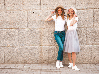 Obraz na płótnie Canvas Portrait of two young beautiful blond smiling hipster girls in trendy summer white t-shirt clothes. Sexy carefree women posing in the street near wall. Positive models having fun in sunglasses and hat