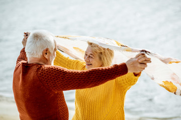 Lovely senior couple playing with scarf during the windy weather, standing together on the sandy...