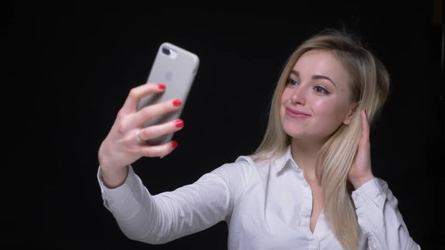 Young and beautiful businesswoman in white shirt making selfies on smartphone on black background.