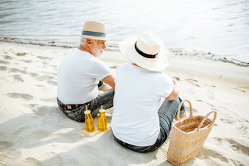 Senior couple sitting together with drinks and bag on the sandy beach, enjoying their retirement near the sea, rear view