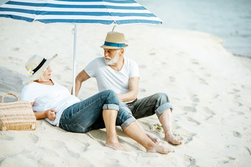 Happy senior couple relaxing, lying together under umbrella on the sandy beach, enjoying their retirement near the sea