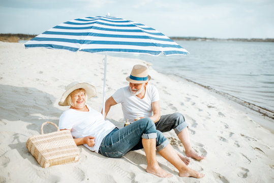 Happy senior couple relaxing, lying together under umbrella on the sandy beach, enjoying their retirement near the sea