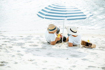 Senior couple sitting together under umbrella on the sandy beach, enjoying their retirement near...