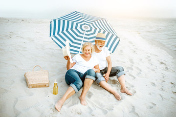 Happy senior couple relaxing, lying together under umbrella on the sandy beach, enjoying their retirement near the sea
