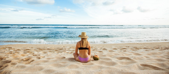 A girl in a hat sits on the white sand with his back to the camera and looks at the ocean, next to it is a coconut. Banner Panorama.