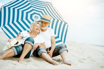 Happy senior couple relaxing, lying together under umbrella on the sandy beach, enjoying their retirement near the sea