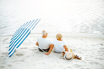 Lovely senior couple relaxing, sitting together with sun umbrella on the sandy beach, enjoying...