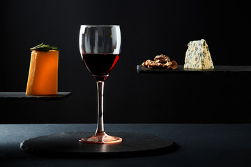 Red wine in crystal glass beside pieces of cheese and walnut on black background