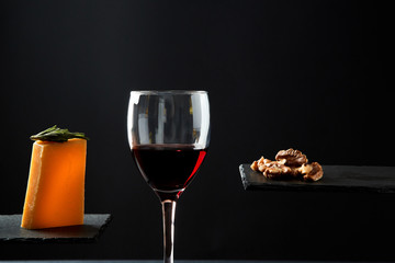 Red wine in crystal glass beside pieces of cheese and walnut on black background
