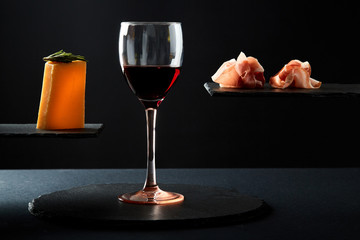Composition of red wine in glass and pieces of prosciutto and cheese snack on black background