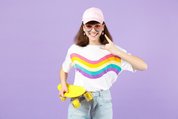 Pretty teen girl in vivid clothes, eyeglasses holding yellow skateboard, showing horns up gesture isolated on violet pastel background. People sincere emotions, lifestyle concept. Mock up copy space.