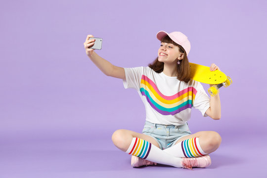 Charming teen girl in vivid clothes holding yellow skateboard, doing selfie shot on mobile phone isolated on violet pastel background. People sincere emotions, lifestyle concept. Mock up copy space.