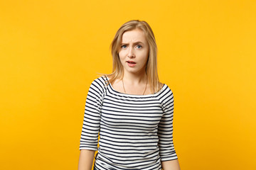 Portrait of perplexed puzzled young woman in striped clothes standing looking camera isolated on yellow orange wall background in studio. People sincere emotions lifestyle concept. Mock up copy space.