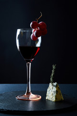 Glass of delicious red wine garnished with bunch of grapes and piece of blue cheese on black background.