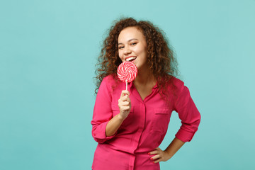 Portrait of cute african girl in casual clothes hold in hand eating pink round lollipop isolated on...