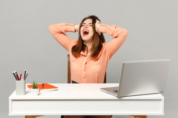 Young crazy screaming woman keeping eyes closed, putting hands on head sit, work at desk with pc laptop isolated on gray background. Achievement business career lifestyle concept. Mock up copy space.