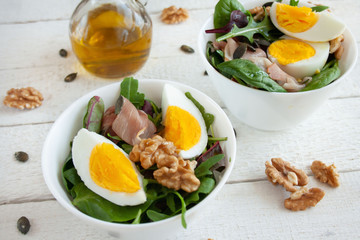 Salad with egg, walnuts and parma ham in white bowls, olive oil in the background
