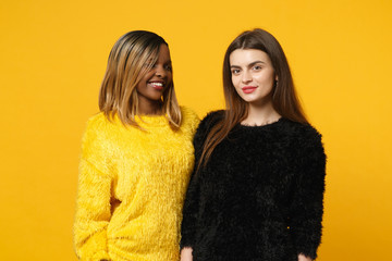 Two young women friends european and african american in black yellow clothes standing posing isolated on bright orange wall background, studio portrait. People lifestyle concept. Mock up copy space.