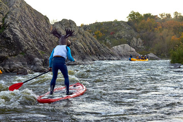 Funny man dressed as an elk (moose) is paddling on a SUP board on fast mountain whitewater river among the rapids at dusk at dusk. Costume water show. Stand up paddle boarding - active recreation.