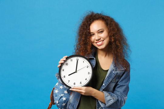 Young african american girl teen student in denim clothes, backpack hold clock isolated on blue wall background studio portrait. Education in high school university college concept. Mock up copy space