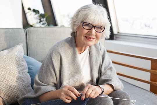 People, retirement, hobby and maturity concept. Indoor image of middle aged positive woman pensioner with gray hair spending winter day at home, knitting scarf for her son, enjoying creative process