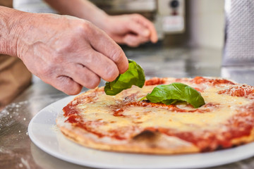 Chef making pizza at Italian restaurant, adding basil, closeup on hands and pizza