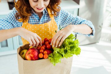 happy woman in polka dot yellow apron taking fruits and vegetables out of paper bag