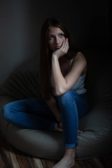 A teen girl sits on a bean bag looking away, in a dark moody setting.