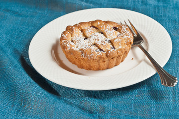 top view, close distance of a freshly baked, homemade, blackberry short bread tart with sprinkles powdered sugar on top, on a round, white plate with an artisan fork on a blue place mat