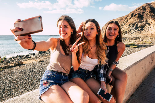Millennial girls taking selfie picture with modern phone for social media having lot of fun together in friendship - smile and crazy expression people outdoor at the beach for summer holiday