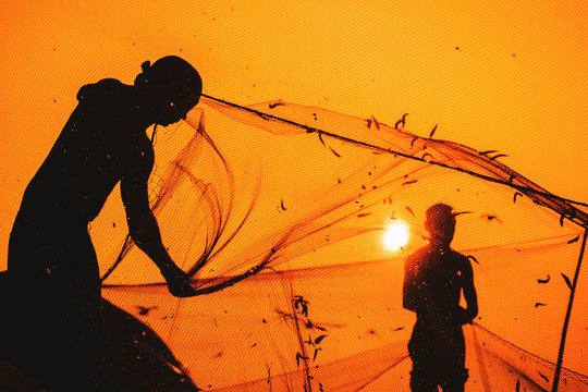 Anonymous, unrecognizable, silhouetted fisherman on the beach shore using fishing net during beautiful, golden sunset