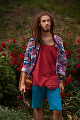 Young handsome bearded man hippie with dreadlocks with his guitar standing on the red roses flower...