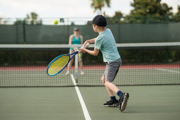 Boy focused with good form playing tennis with mom on holiday