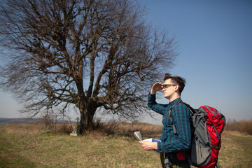 A traveler with a backpack, looking at the map and walking in the countryside. Tree in the background