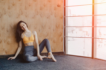 Fototapeta premium Young attractive fitness girl sitting on the floor near the window on the background of a wooden wall, resting on yoga classes