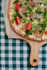 Neapolitan pizza with salami, arugula, tomatoes sprinkled with cheese on a wooden board on a tablecloth in a cell with a place for text