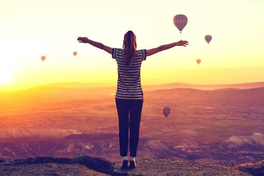 A Woman Or Girl Admires A Beautiful View Of Hot Air Balloons In Cappadocia In Turkey And Raises Her Arms Up.