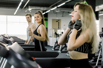 Young sports friends making cardio workout on stationary treadmill in the gym. Two girls and their handsome trainer running in machine treadmill at fitness gym club