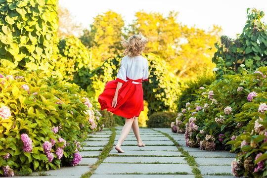 Full Length Back View Of Happy Careless Barefoot Attractive Young Blonde Woman In Stylish Red White Dress Walking With Happiness On Tile Path In Garden. Outdoor Shot In The Park At Summer Daytime.