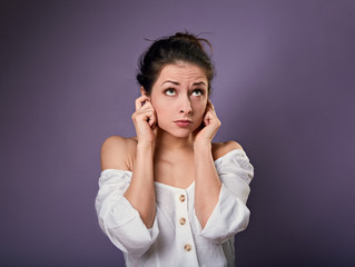 Stressed uncertain casual woman closed ears the fingers because not want the hear any sounds and noise looking up on purple background.