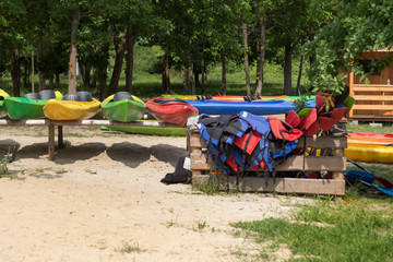 Wypożyczalnia kajaków. Kolorowe kajaki poustawiane na stojakach leżą na plaży. Kapoki i wiosła poukładane w drewnianym pojemniku. 