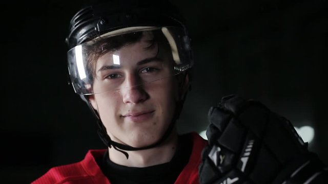 Smiling at camera on dark arena, close up portrait of defender or forward canadian player. CU Portrait of Caucasian male ice hockey player in red uniform, looking into the camera.
