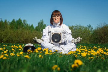 Beautiful girl astronaut without a helmet sits on a green lawn among flowers in a meditative...