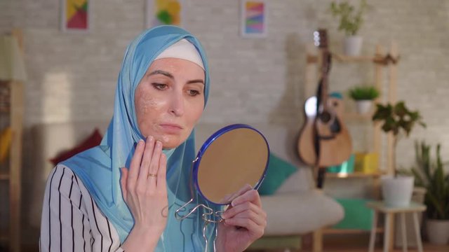 Muslim woman in a hijab looks in the mirror at her scar from a burn on her face and cries
