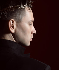 Young man in crown of thorns, martyr and black suit jacke stands back to us with turned head and...